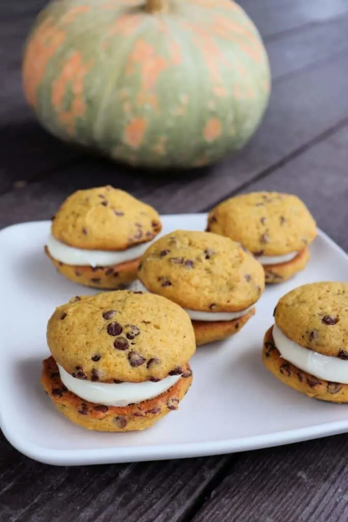 A plate full of sandwich cookies with chocolate chips on a table with a pumpkin in the background.