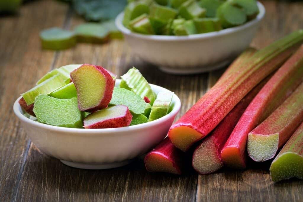 White bowls full of chopped rhubarb pieces sitting next to stalks of uncut rhubarb on a cutting board.