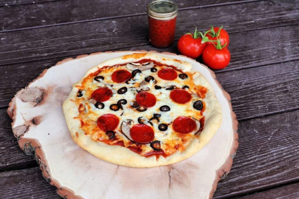 A cooked pizza sitting on top of a round wooden cutting board with fresh tomatoes and a jar of pizza sauce sitting behind it. 