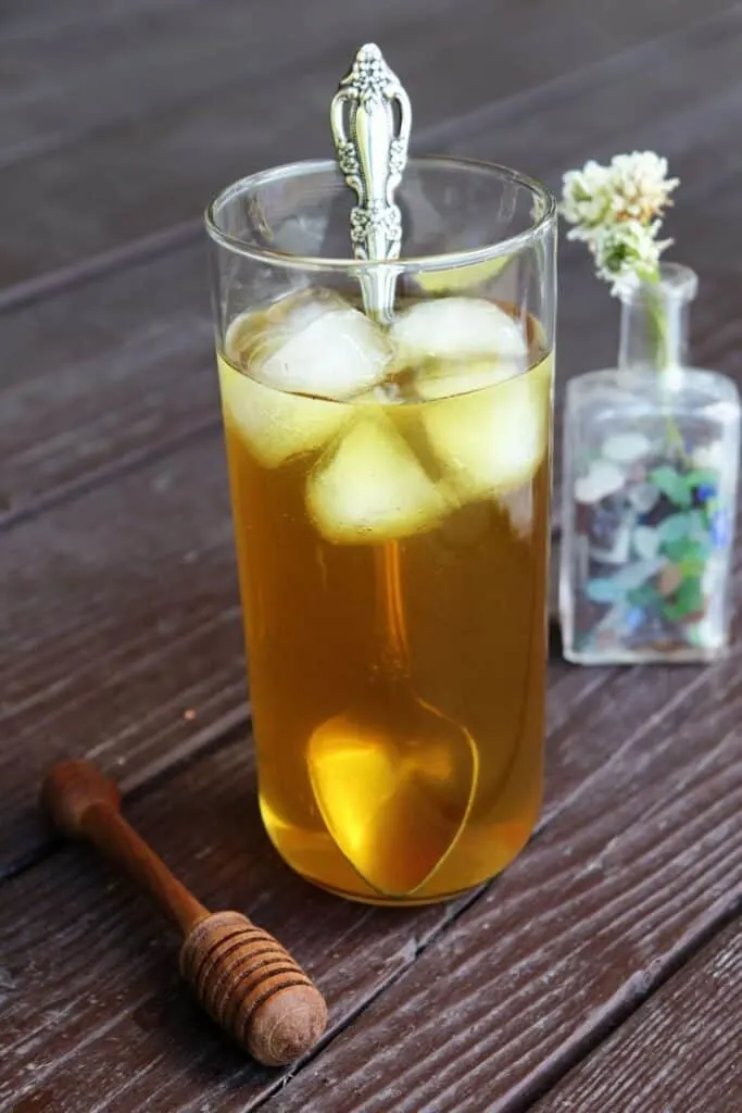 A glass full of ice and white clover tea with a long silver spoon sticking out of it, a wooden honey dipper sitting to the left, a small bottle full of white clovers in the background.
