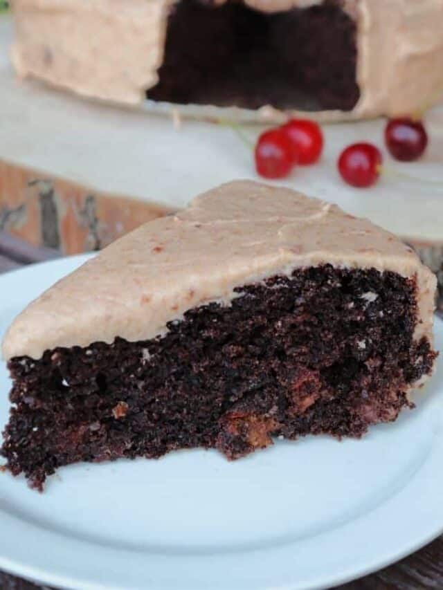 Close up of a slice of frosted chocolate cake on a white plate with the remaining cake sitting on a round wooden plank with fresh cherries behind it.