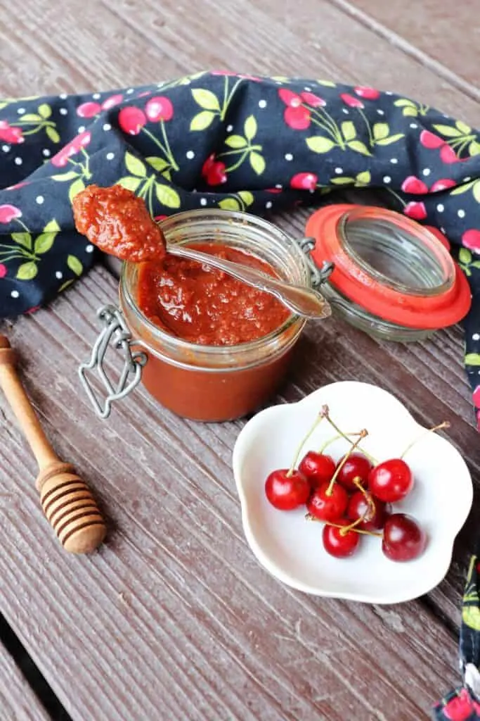 An open jar of sour cherry butter as seen from above with a spoon sitting on top of it, surrounded by fresh cherries in a bowl, a honey dipper, and black cloth with cherries decorating it.
