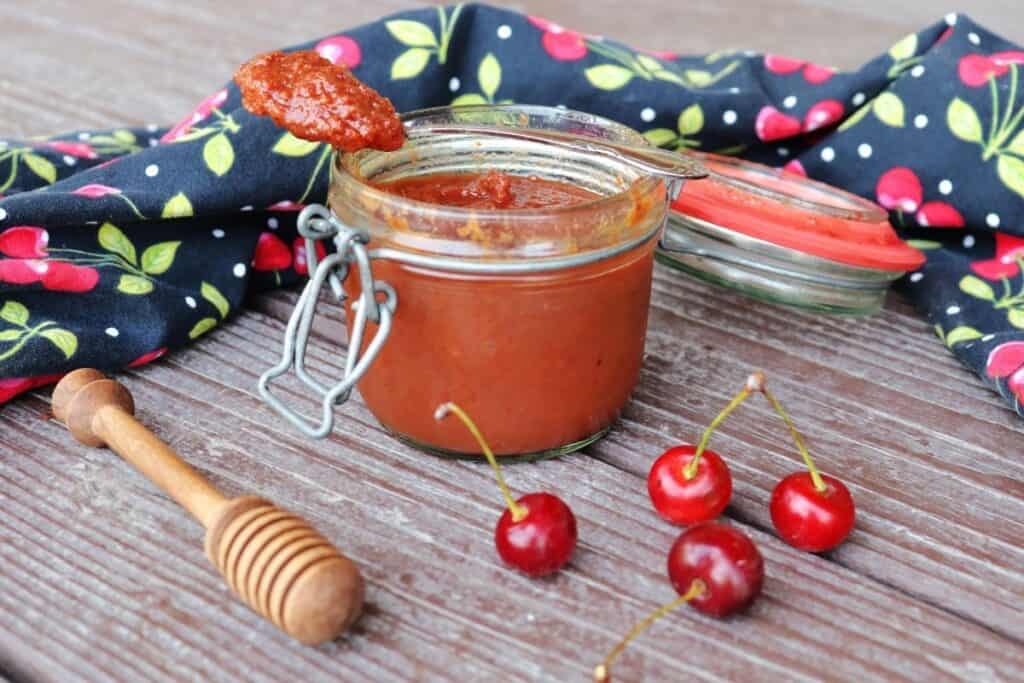 An open jar of sour cherry butter with a spoon sitting on top of it, surrounded by fresh cherries, a honey dipper, and black cloth with cherries decorating it.