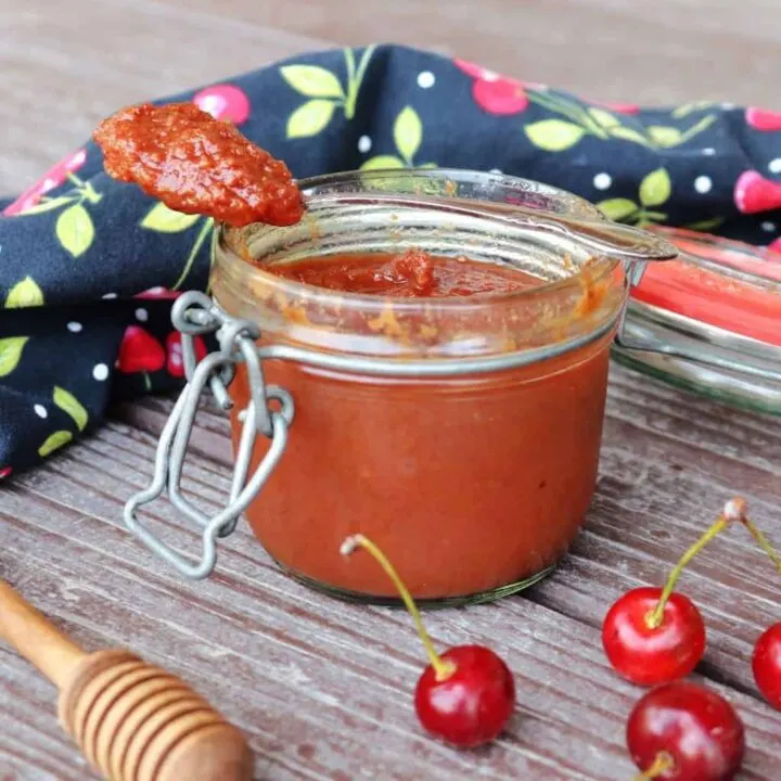 An open jar of sour cherry butter with a spoon sitting on top of it, surrounded by fresh cherries, a honey dipper, and black cloth with cherries decorating it.