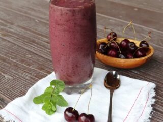 A glass full of cherry smoothie sitting on a white napkin with a spoon, fresh cherries, and sprig of mint.
