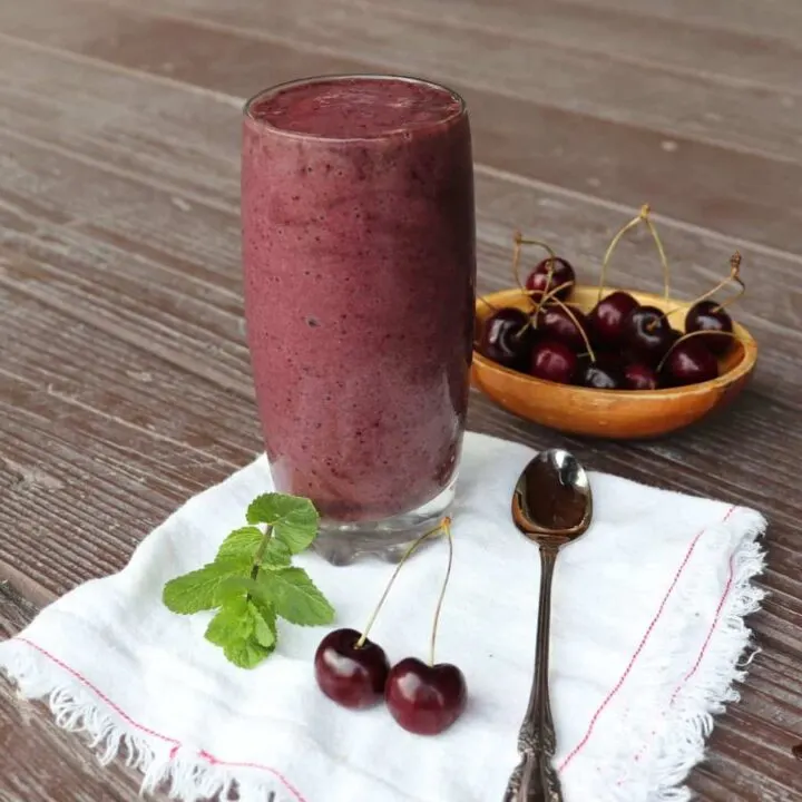 A glass full of cherry smoothie sitting on a white napkin with a spoon, fresh cherries, and sprig of mint.