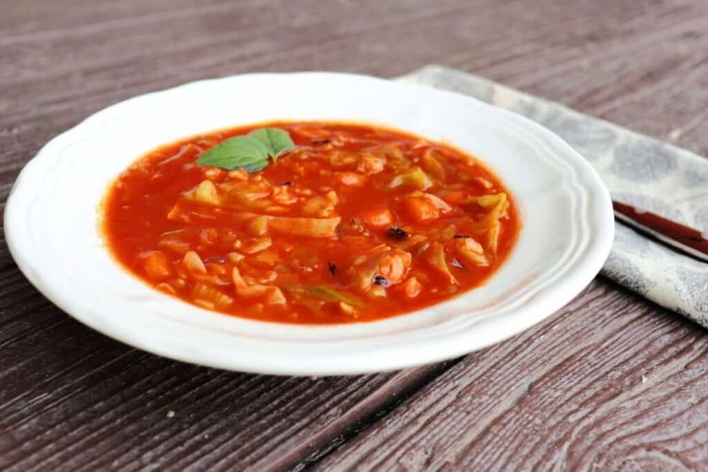 A white bowl full of cabbage roll soup as seen from the side with a napkin and spoon sitting to the right. 