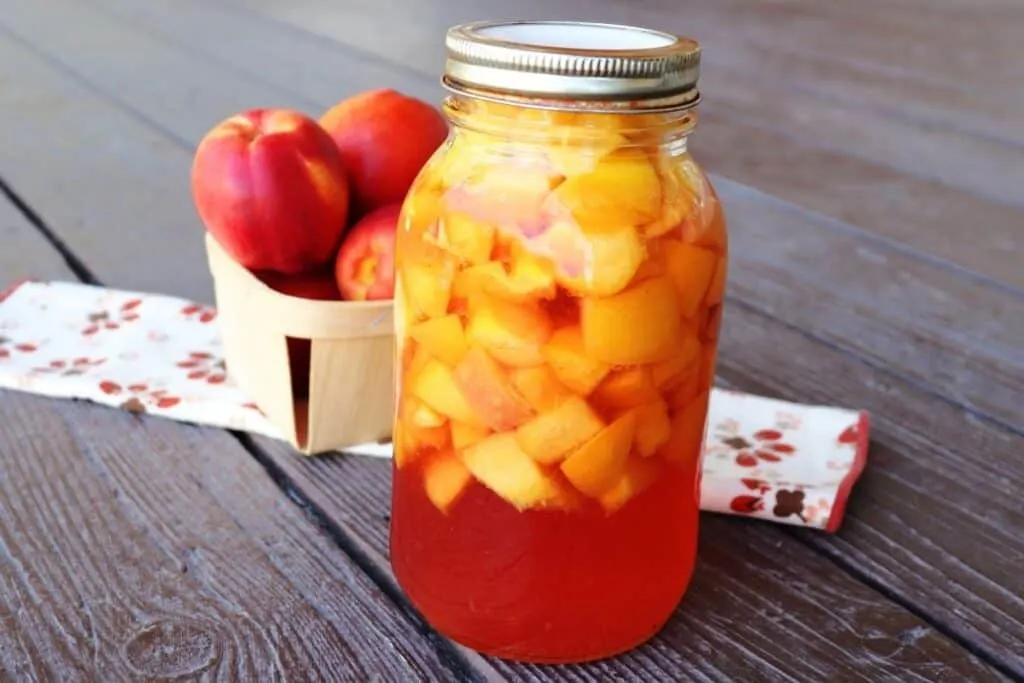 A jar of canned nectarines sitting in front of a cloth table runner with a basket of fresh nectarines sitting on it. 