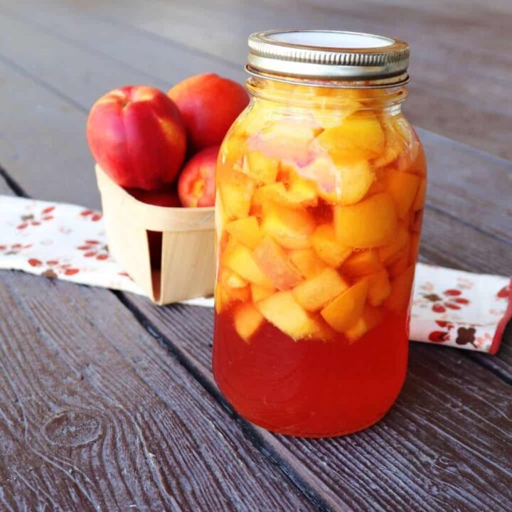 A jar of canned nectarines sitting in front of a cloth table runner with a basket of fresh nectarines sitting on it.