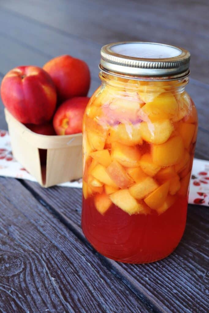 A jar of canned nectarines sitting in front of a cloth table runner with a basket of fresh nectarines sitting on it. 