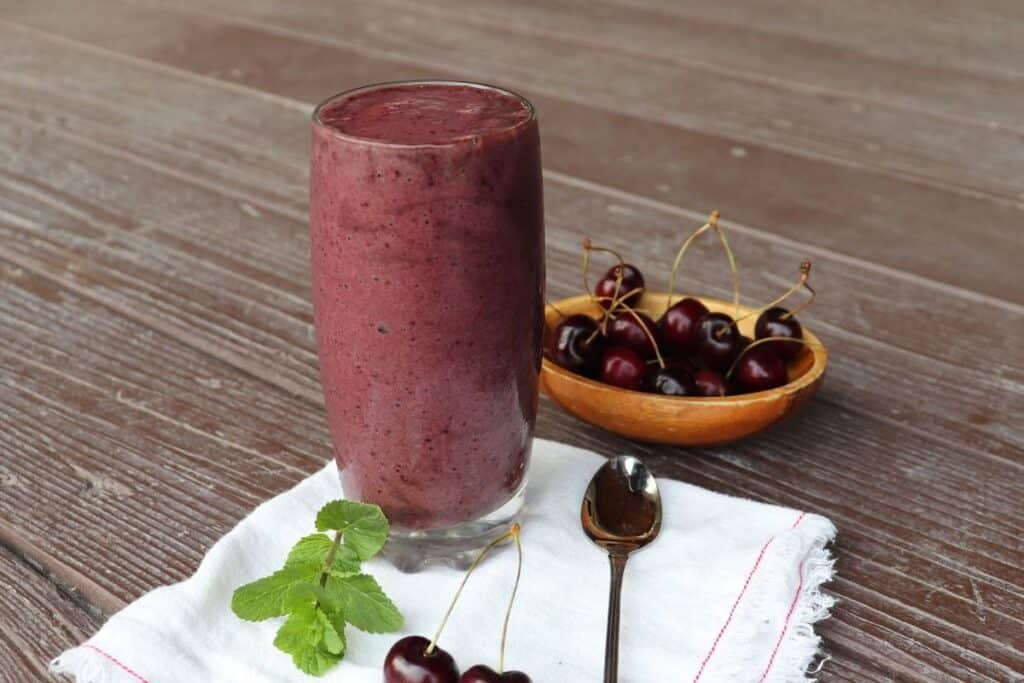 A glass full of cherry smoothie sitting on a white napkin with a spoon, fresh cherries, and a sprig of mint, a wooden bowl of fresh cherries in the background.