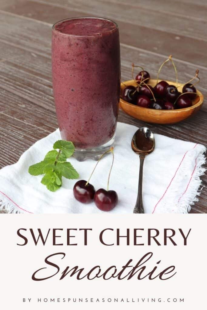 A glass full of cherry smoothie sitting on a white napkin with a spoon, fresh cherries, and a sprig of mint, a wooden bowl of fresh cherries in the background with text overlay reading: Sweet Cherry Smoothie.