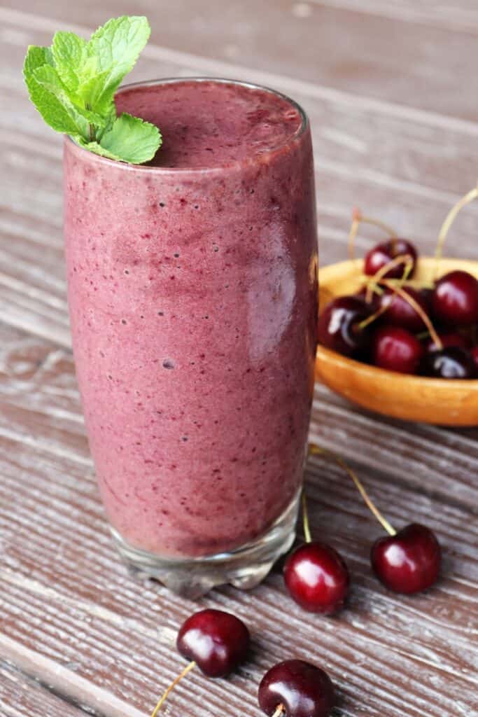 A drinking glass full of cherry smoothie with a sprig of fresh mint on top surrounded by fresh cherries.