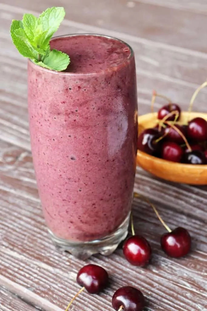 A drinking glass full of cherry smoothie with a sprig of fresh mint on top surrounded by fresh cherries.