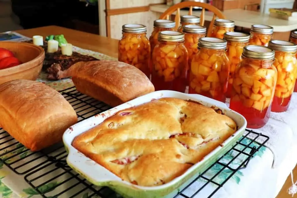A kitchen table full of quart jars of canned nectarines, 2 loaves of bread on a cooling rack, and a pan of cobbler on a cooling rack. 