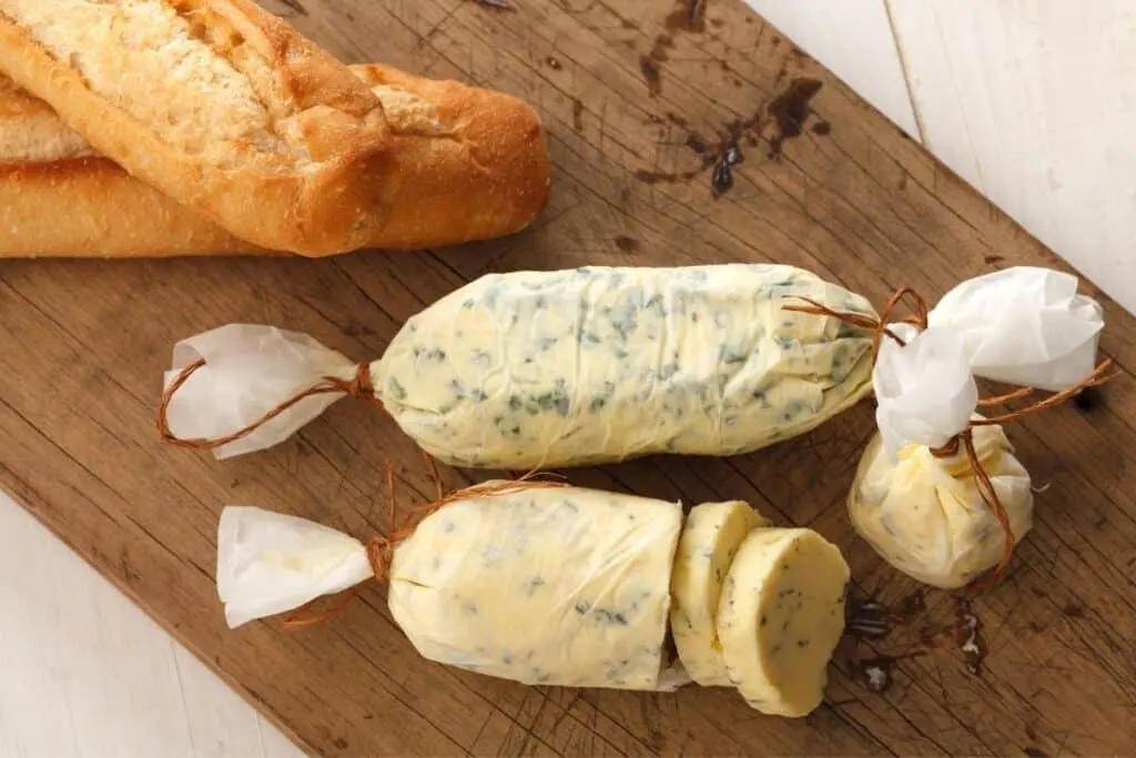 Two round logs of compound butter wrapped in wax paper sitting on a wooden board with loaves of bread.