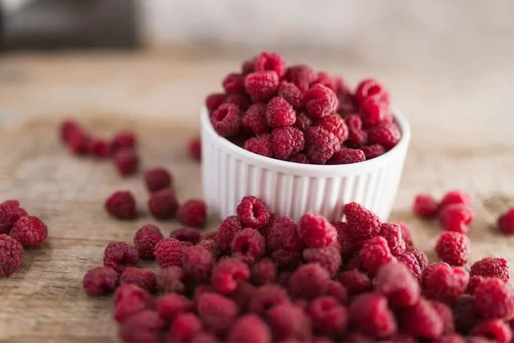 A white bowl of fresh raspberries sits on a table surrounded by more red raspberries.
