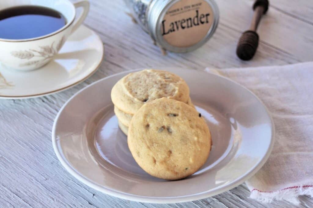 White Chocolate Lavender Cookies stacked on a plate with a cup of tea, jar of lavender, and wooden honey dipper in the background. 