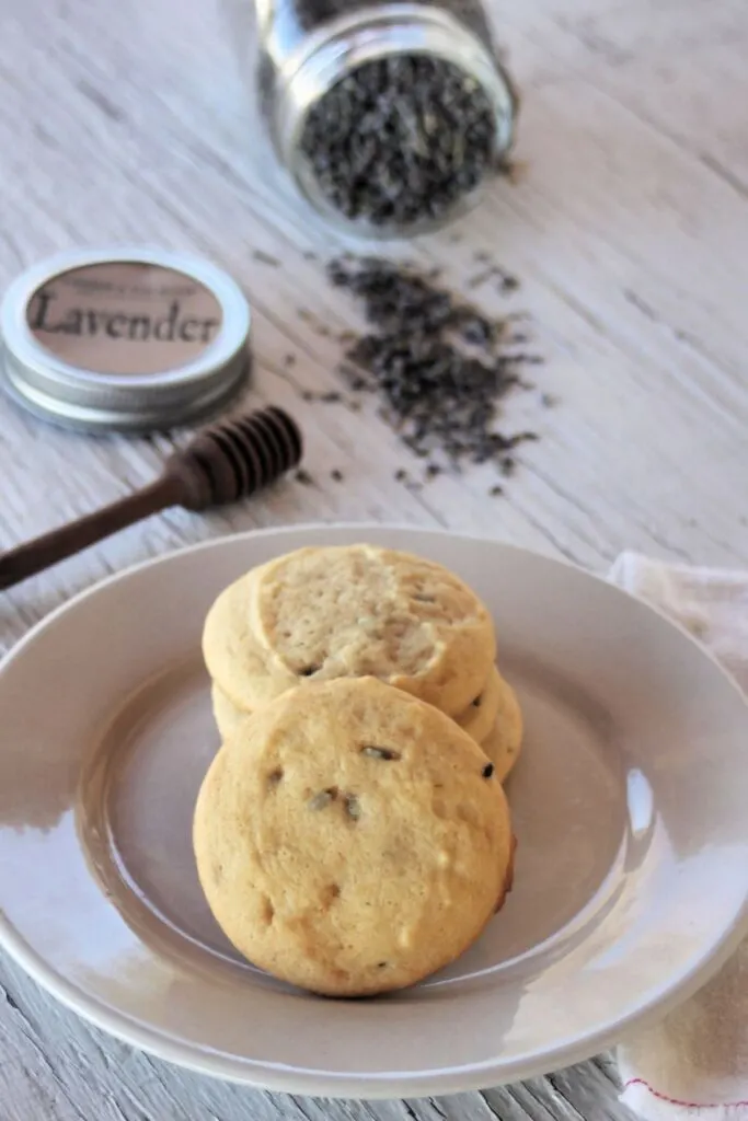 White chocolate lavender cookies stacked on a white plat with a jar of lavender in the background and lavender spilling onto the table.