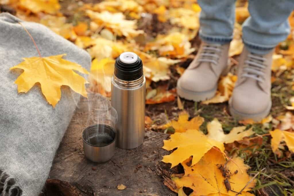 A thermal bottle sitting next to the full cap on a tree trunk surrounded by yellow fall leaves and the boots of  person standing on the ground. 