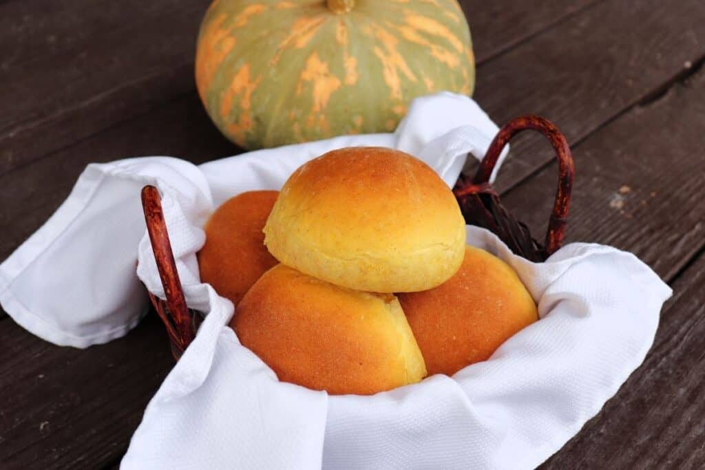Pumpkin dinner rolls in a white linen lined basket with a pumpkin behind it.