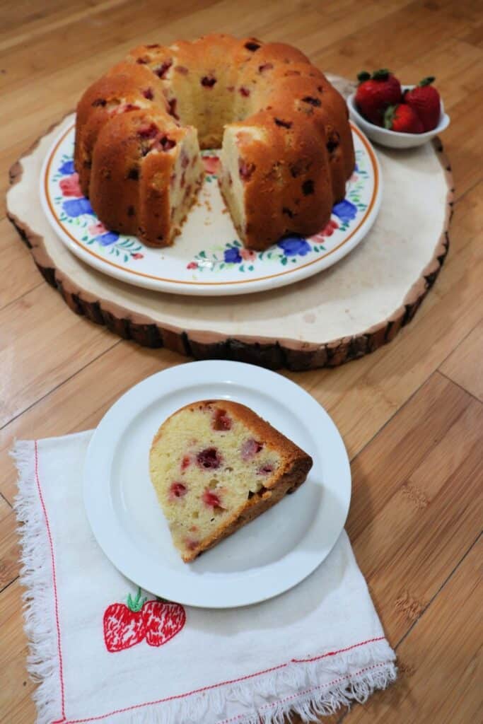 A slice of strawberry bundt cake sitting on a white plate, sitting on a napkin with embroidered strawberries on it. The remaining cake and bowl of strawberries in the background.