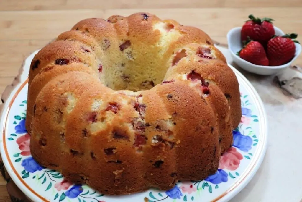 A strawberry bundt cake sitting on a floral cake plate with a bowl of fresh strawberries in the background.