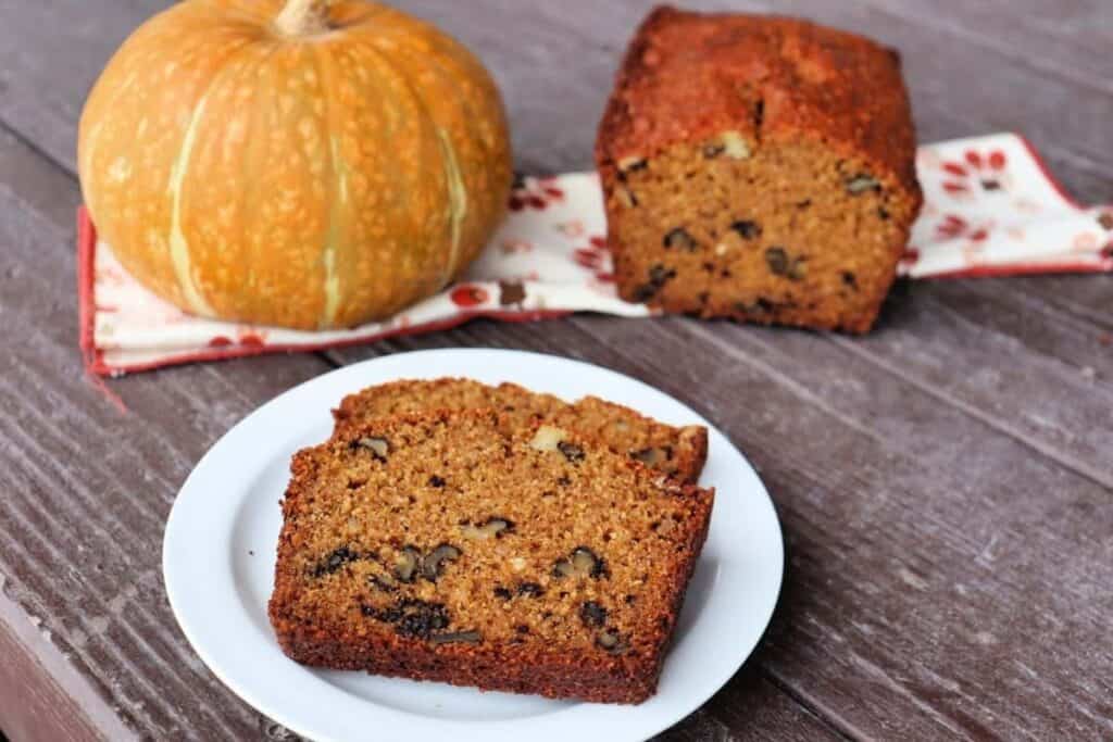 slices of pumpkin walnut bread sit on a plate. In the background is the remaining loaf and a pumpkin sitting on a floral napkin.