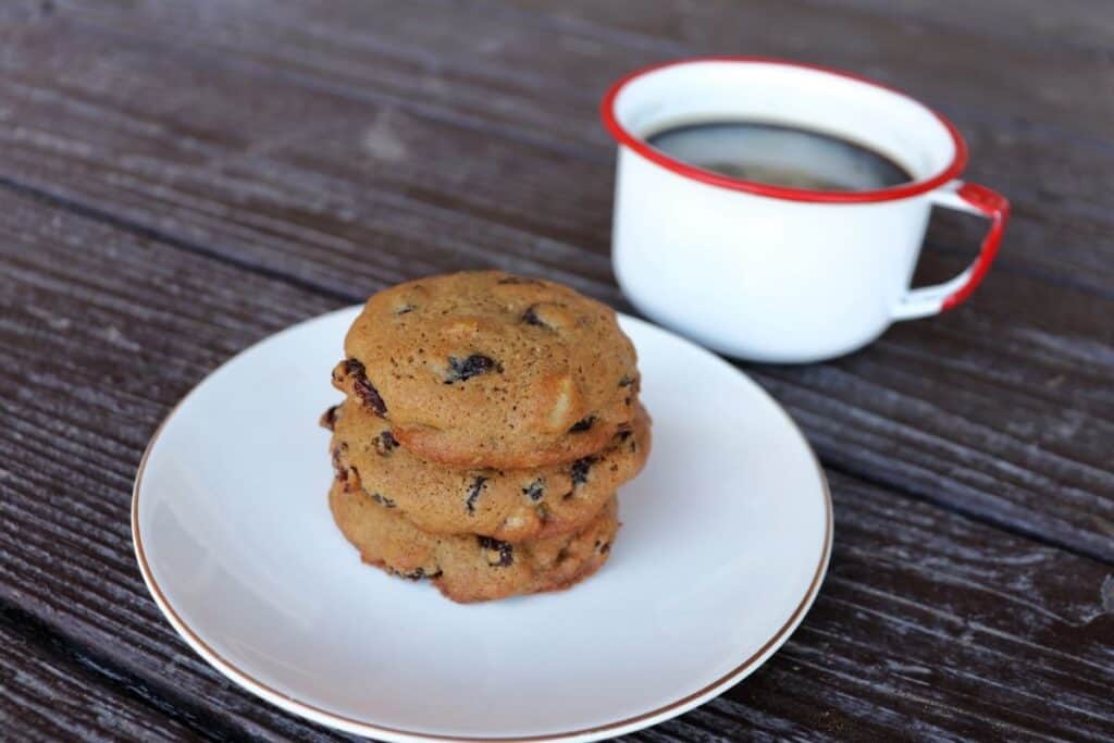 A stack of 3 cookies on a plate with a cup of coffee in the background.