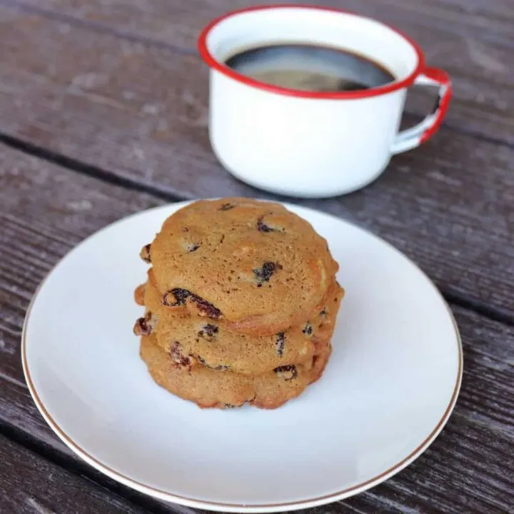 A stack of 3 cookies on a plate with a cup of coffee in the background.