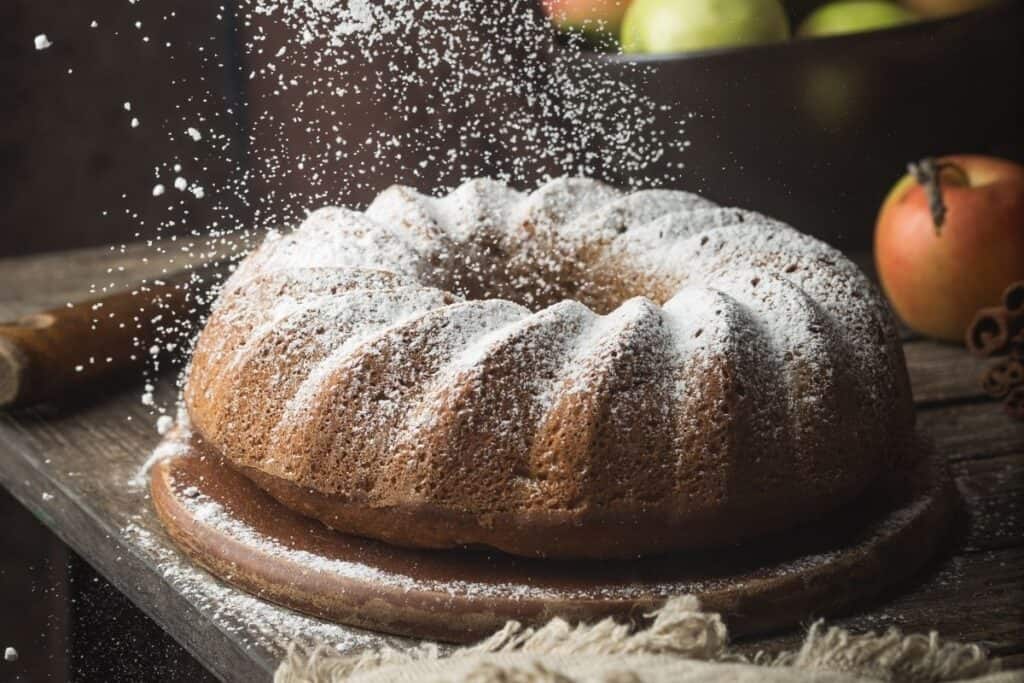 A bundt cake on a wooden plate being sprinkled with powdered sugar with fresh apples in the background.