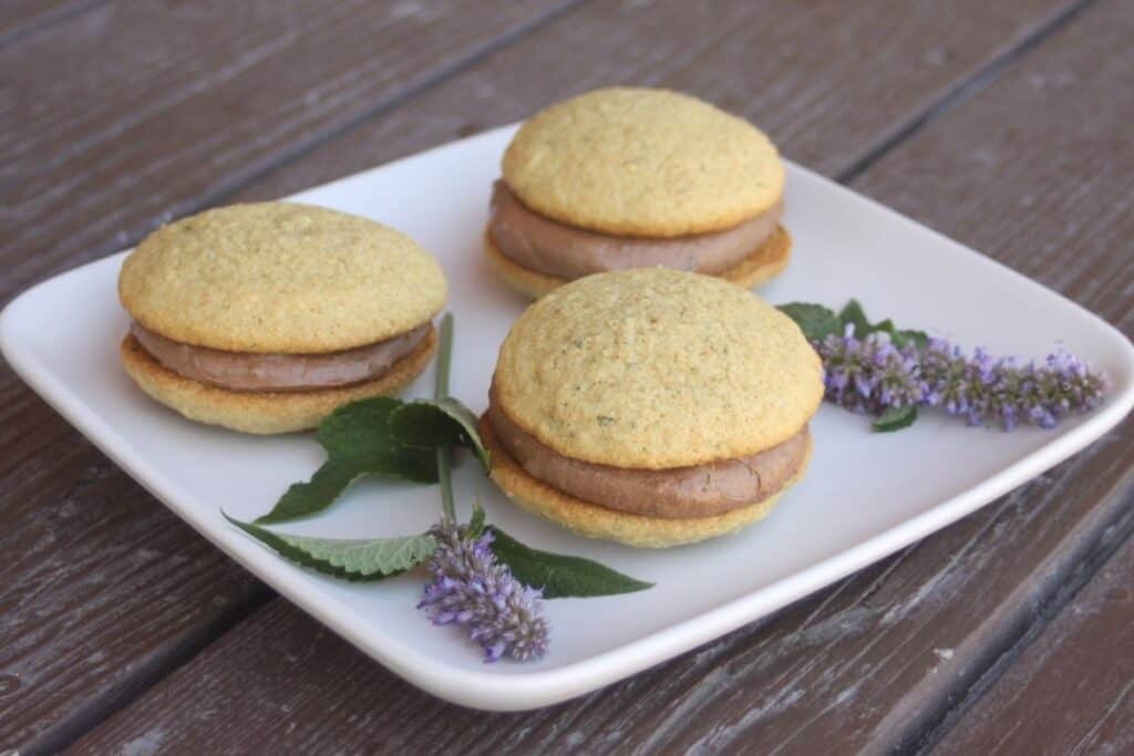 White whoopie sandwiched around a chocolate filling on a white plate with fresh stems of anise hyssop flowers. 