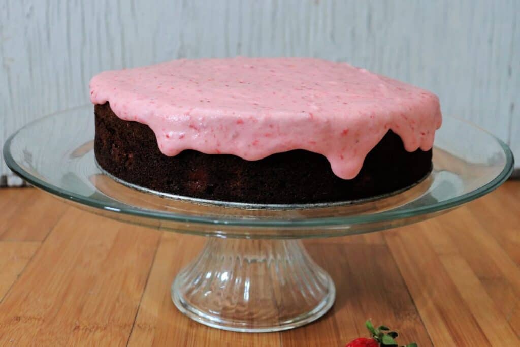 A chocolate cake with pink frosting dripping down the sides sits on a glass cake plate.