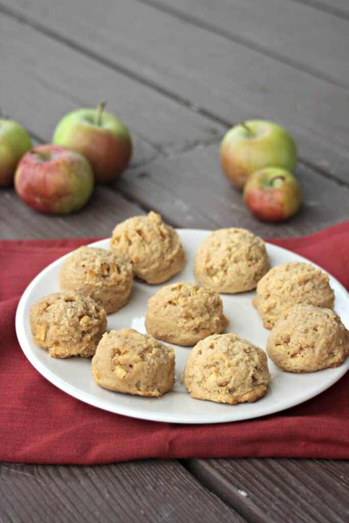 Apple peanut butter cookies on a plate sitting on top of a red cloth. Fresh apples scattered on the table behind the plate.