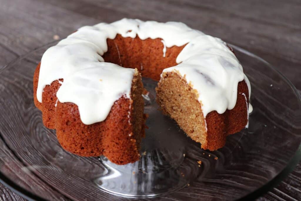 An applesauce bundt cake with a vanilla frosting that has had a slice removed sitting on a glass cake plate.