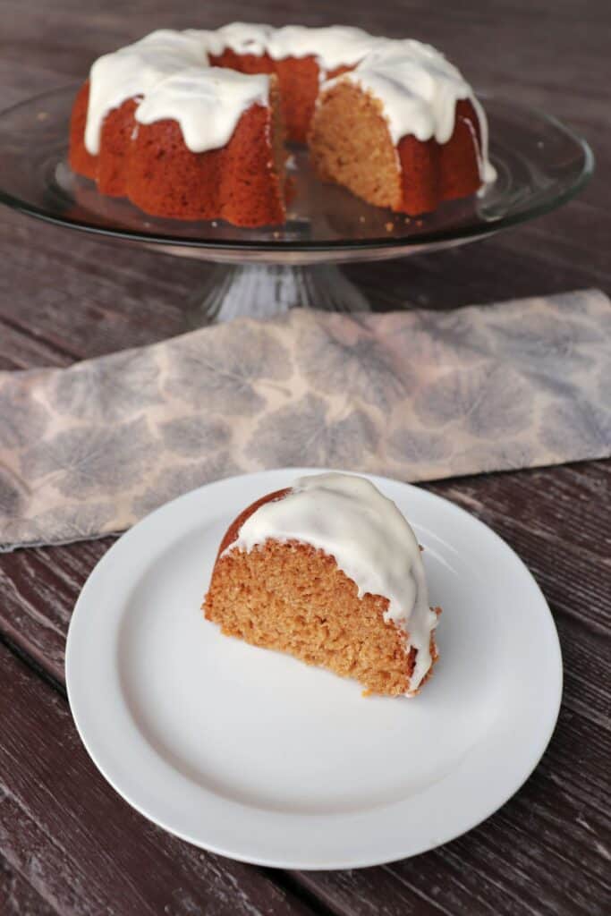 A slice of applesauce bundt cake on a white plate with a napkin sitting behind it. The remaining cake on a glass cake plate in the background.