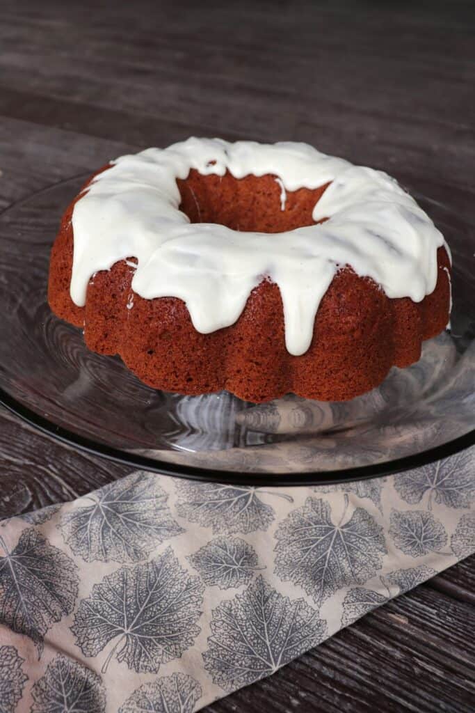 An applesauce bundt cake with vanilla glaze sitting on a glass cake plate with a napkin on the table.