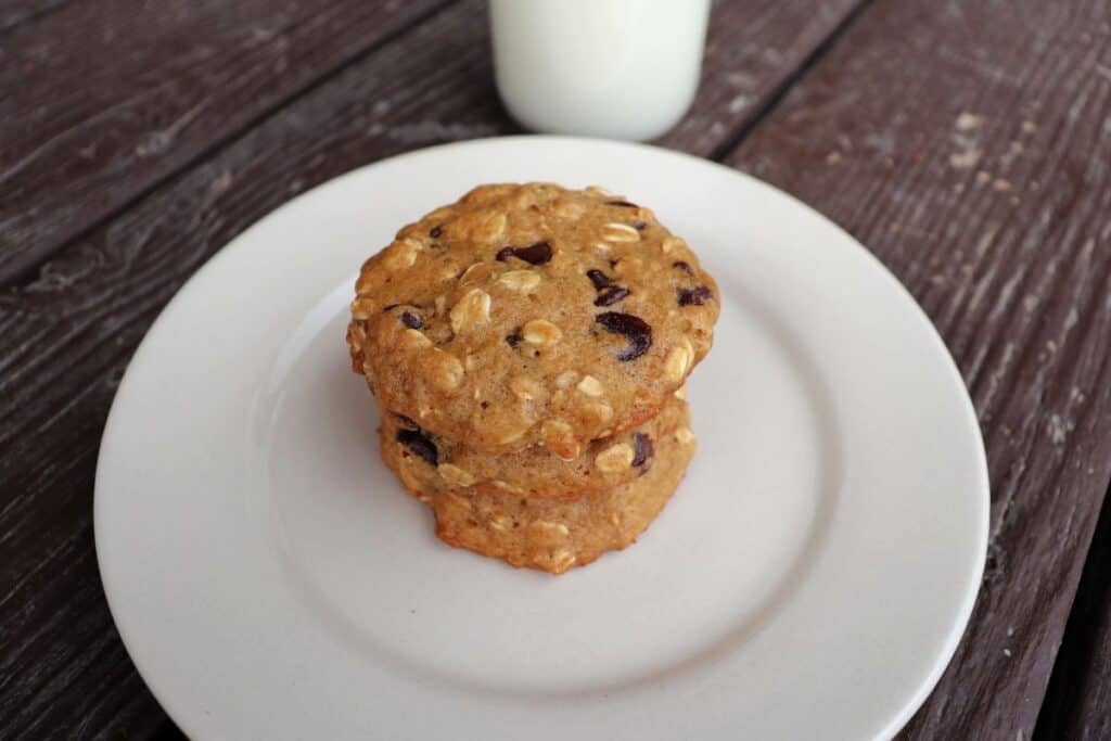 A close-up of 3 applesauce chocolate chip cookies stacked on a plate with a glass of milk in the background.