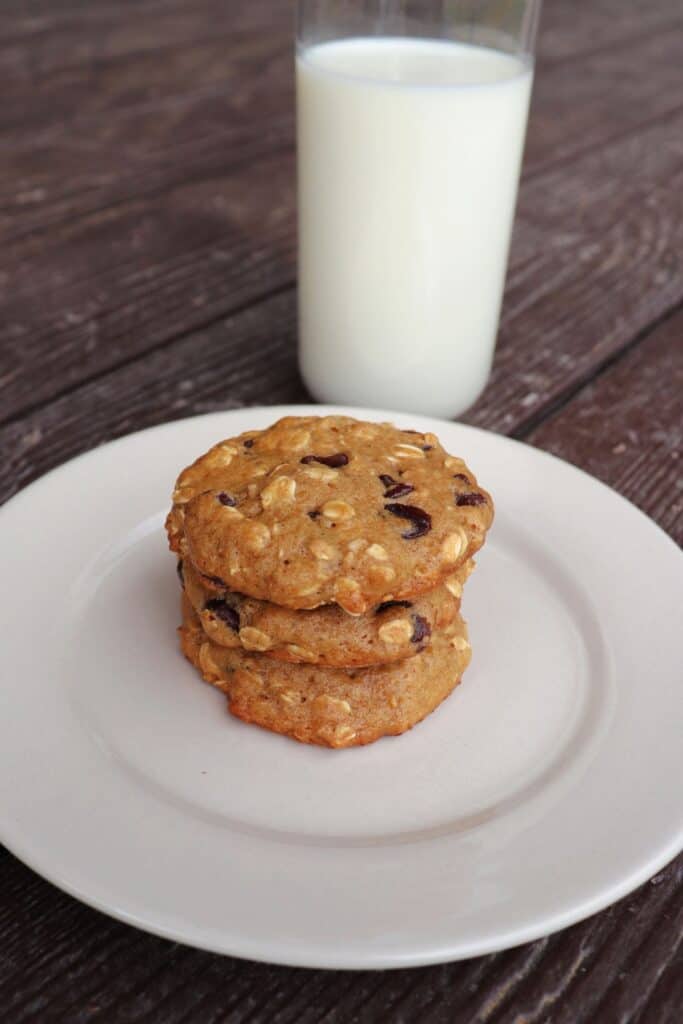 3 applesauce chocolate chip cookies stacked up on each other on a plate, a glass of milk in the background.
