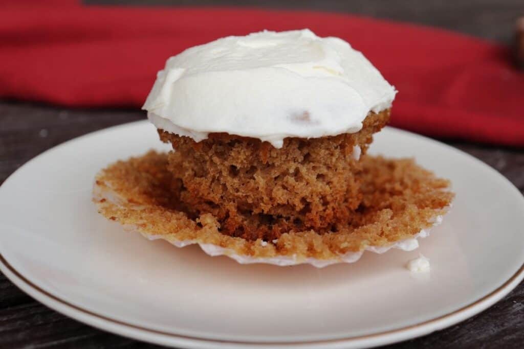 A side view of a frosted applesauce cupcake sitting on top of its paper liner on a white plate with a red cloth in the background.