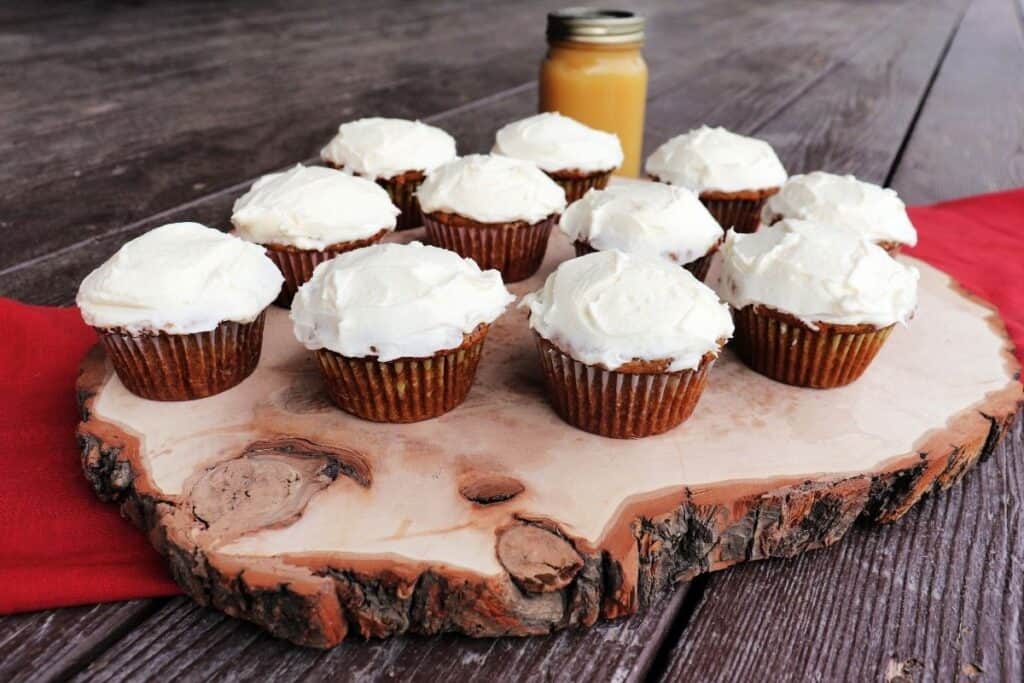 A wooden plank platter scattered with frosted applesauce cupcakes sitting on a red cloth. A jar of applesauce sits behind it.