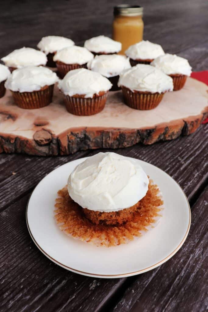 An applesauce cupcake with the paper liner removed sitting on a white plate. A platter of more cupcakes and a jar of applesauce sit in the background.