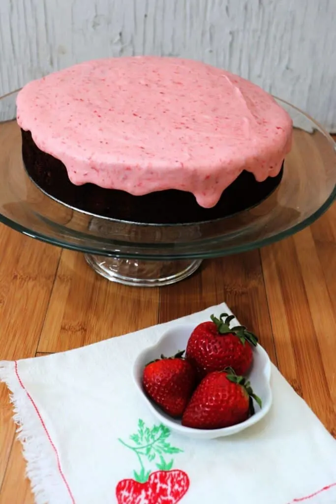 A chocolate cake with pink frosting sits on a glass cake plate with a bowl of strawberries underneath it sitting on a napkin.