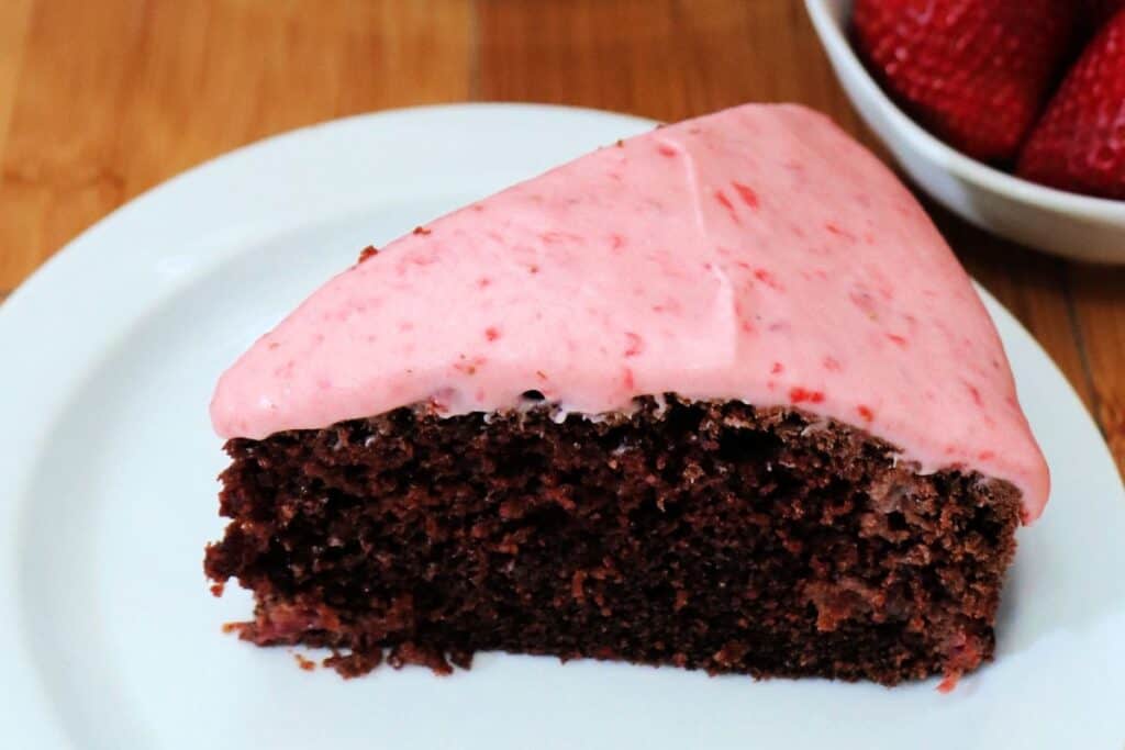 A slice of chocolate cake with strawberry frosting sitting o a white plate.