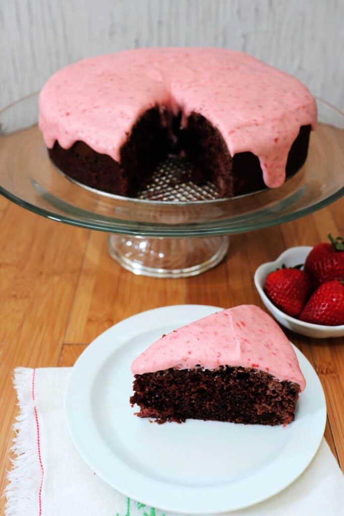 A slice of chocolate cake with pink frosting sits on a white plate, the remaining cake sits on a glass cake plate behind it with a bowl of fresh strawberries.