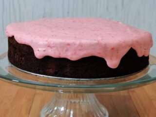 A chocolate cake with pink frosting dripping down the sides sits on a glass cake plate.