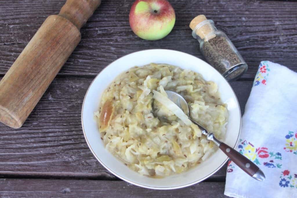 A bowl of cooked sauerkraut and apples with a spoon in it as seen from above. A wooden mallet, fresh apple, jar of seeds, and napkin surround the bowl.