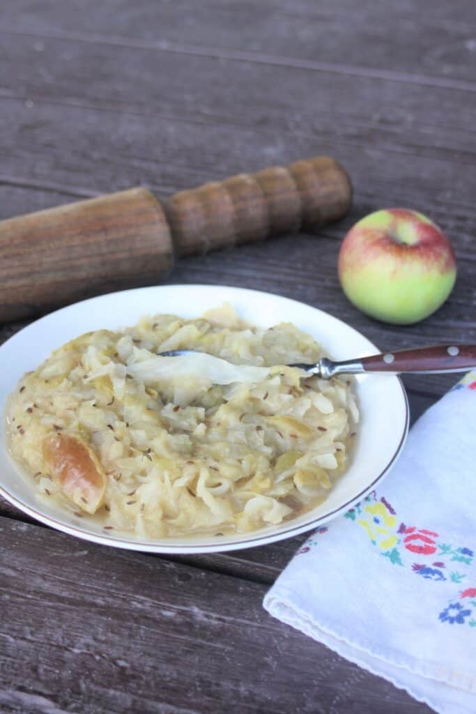 A bowl of cooked sauerkraut and apples with a spoon in it. A wooden mallet, fresh apple,  and napkin surround the bowl.