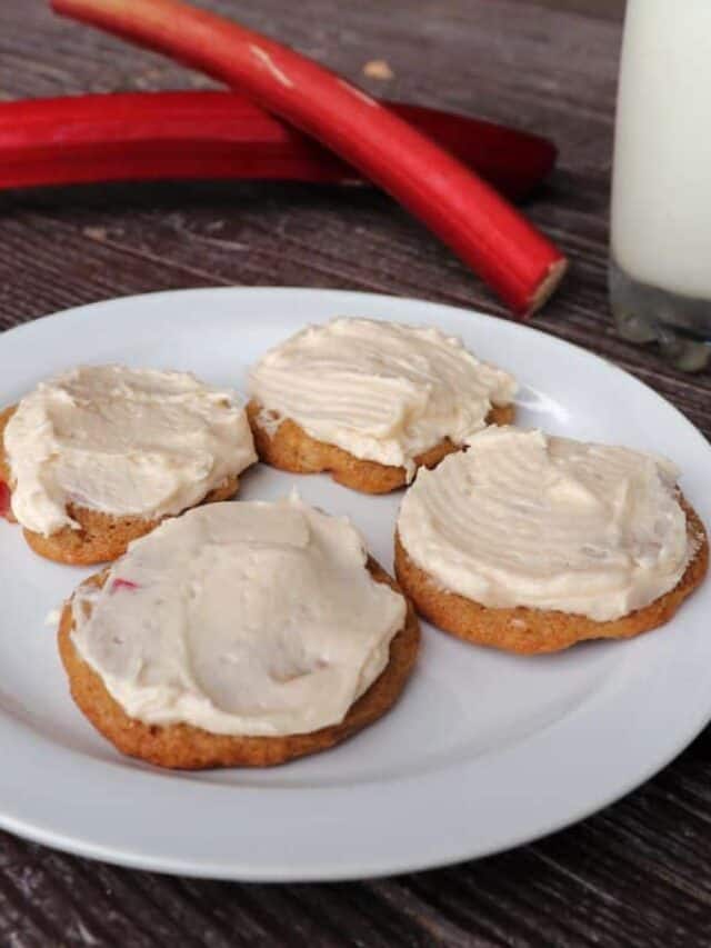 4 frosted rhubarb cookies on a plate. Fresh rhubarb stalks and a glass of milk sit in the background.