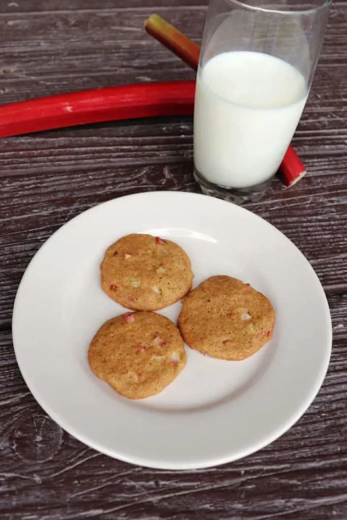 3 rhubarb cookies on a plate with fresh rhubarb stalks and a glass of milk sitting in the background.